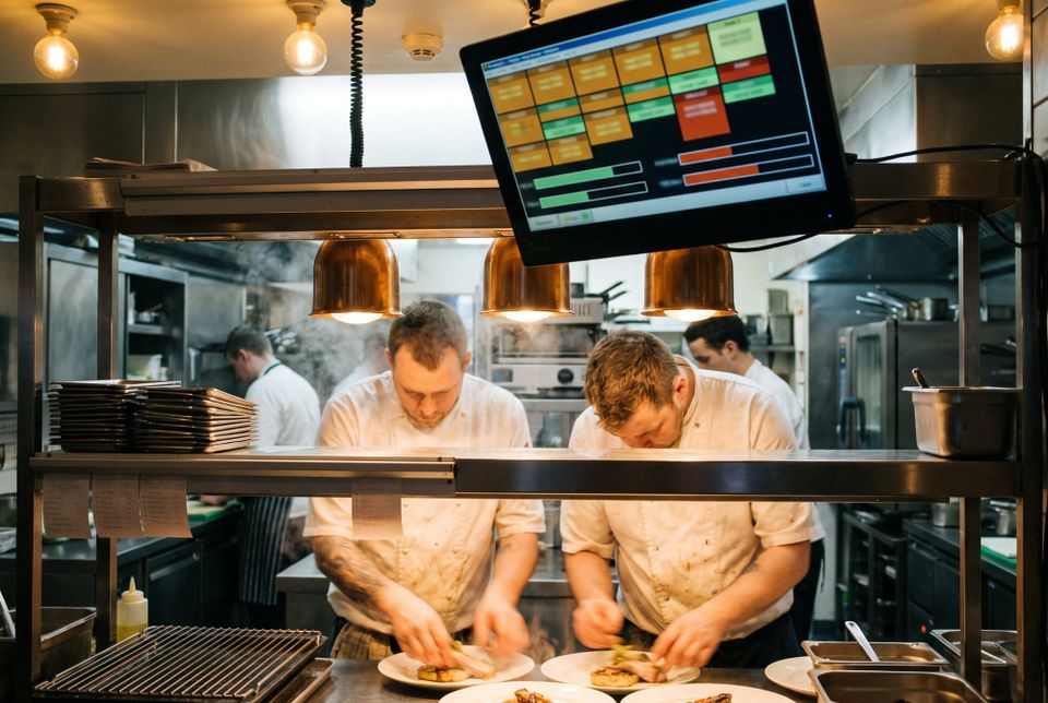 A kitchen display screen above the pass during service, showing colour coded order tickets, with chefs plating dishes underneath beneath copper heat lamps.