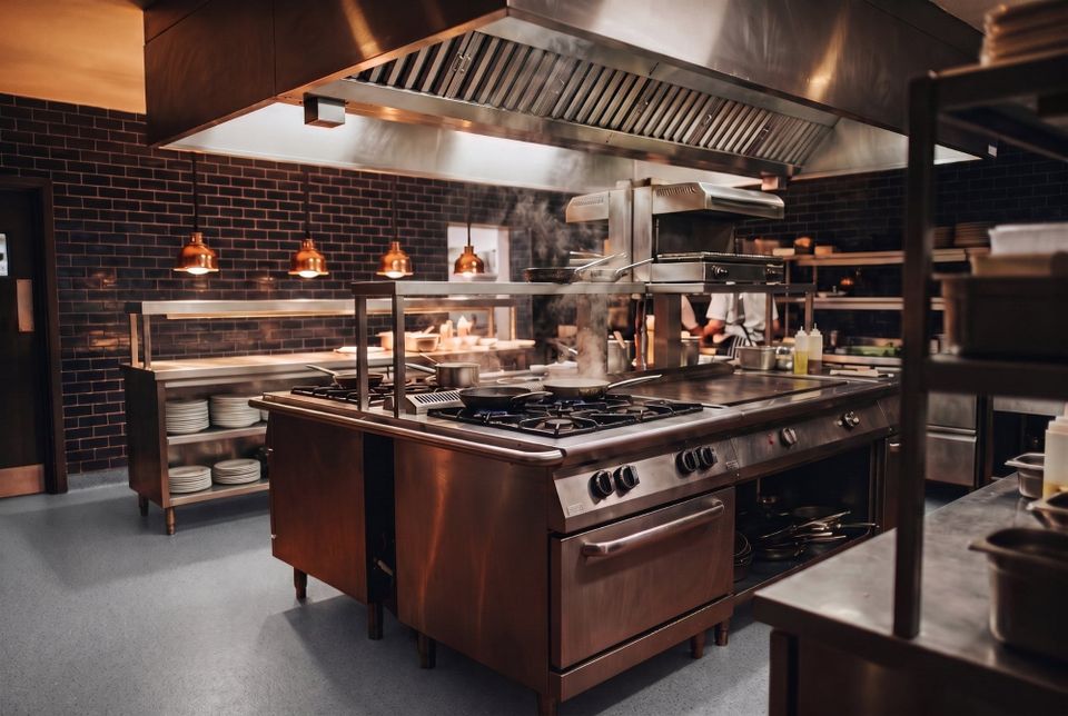 A stainless steel island cooking suite under an extraction canopy in a working commercial kitchen, with a pan steaming on the gas burners and a stainless service pass along the back wall.
