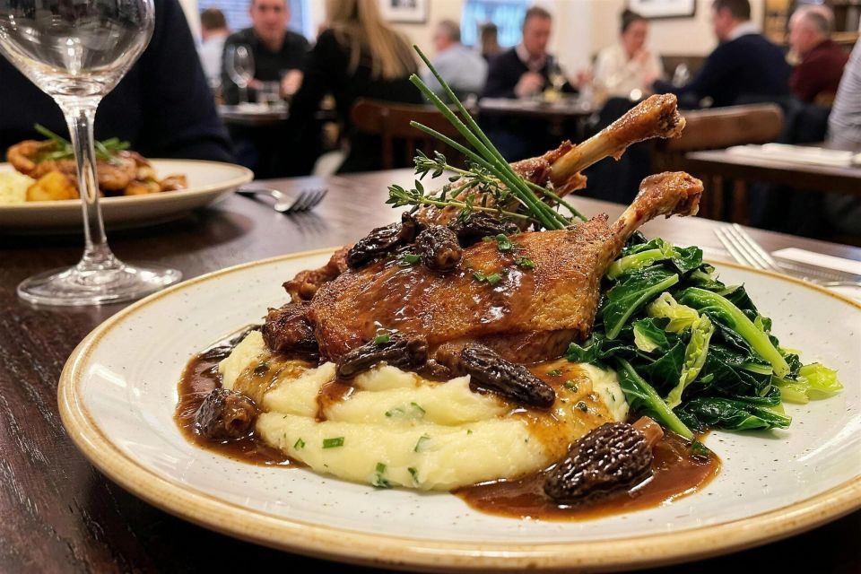 Confit duck leg on mash with greens and morel sauce, plated in a busy restaurant dining room.
