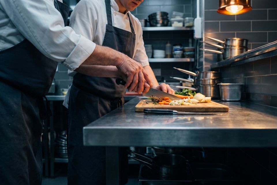 Two chefs at a stainless steel bench in a commercial kitchen, one cutting vegetables on a board while the other works alongside.