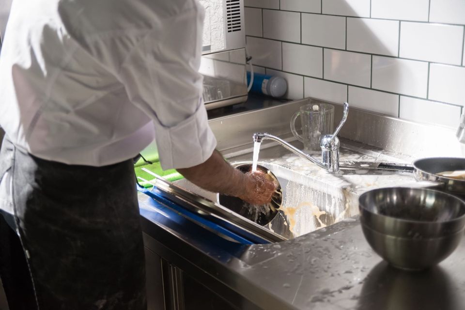 A chef in whites washing a pan under running water at a stainless commercial sink with white tiled walls.