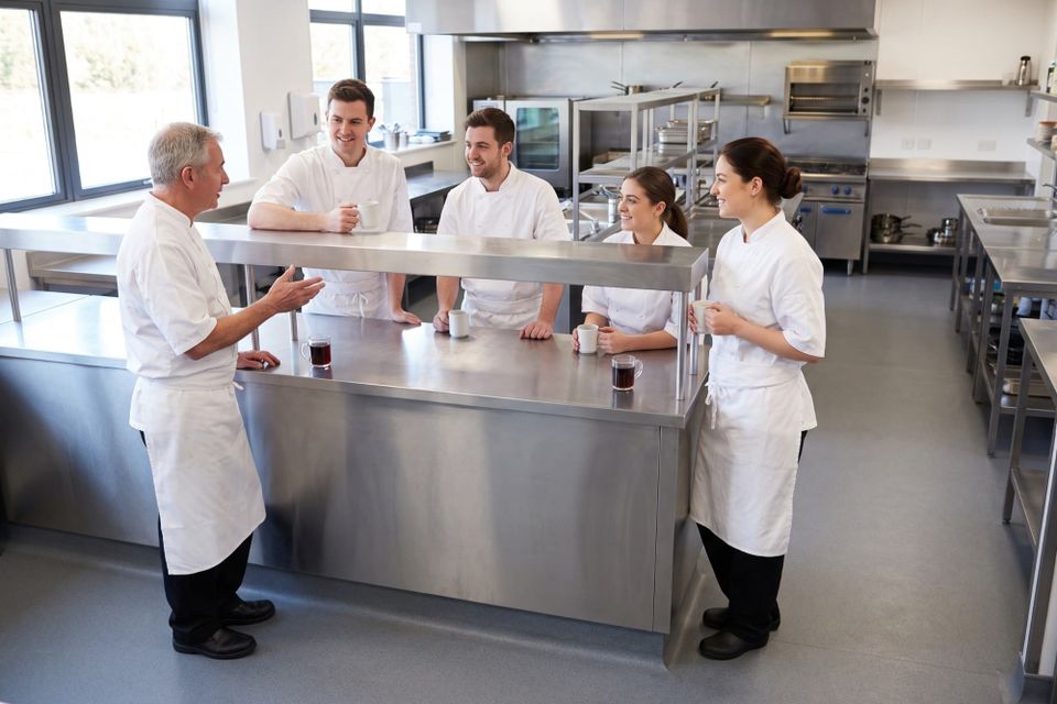 Five kitchen staff in whites gathered round a stainless pass with mugs, one talking and the rest listening.