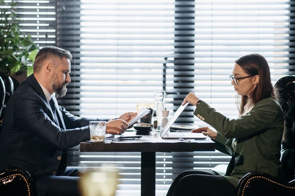 Two people in business dress across a restaurant table, one reading a stapled document, the other closing a laptop.