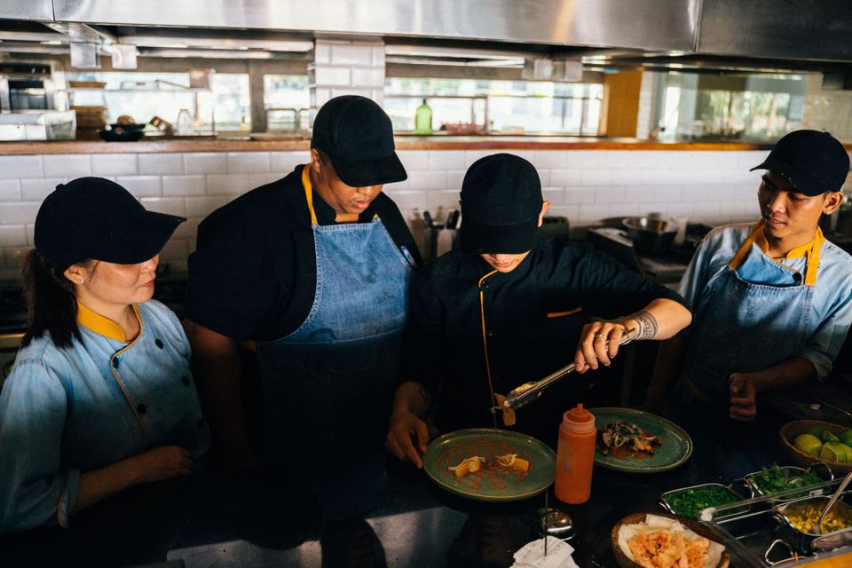 Four kitchen staff in caps and aprons watching a colleague demonstrate with tongs at the stove.