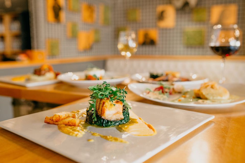 A plated white fish dish with green herbs and a yellow sauce on a wooden pass, more plates waiting behind.