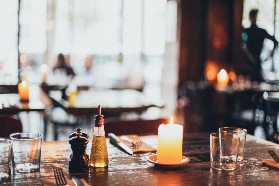 A lit candle on a laid restaurant table with glasses, an oil bottle and a pepper grinder at dusk.