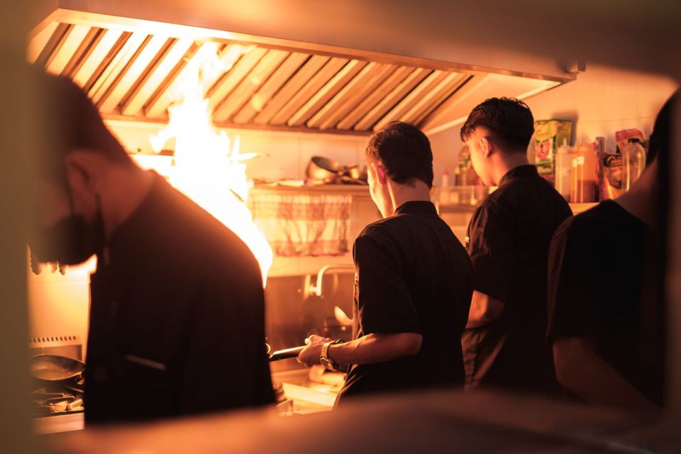 A stainless extraction canopy with baffle filters above a restaurant cook line, flames rising from a pan beneath.