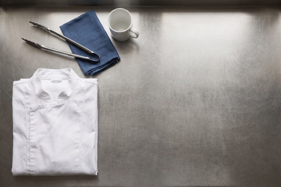 A folded chef jacket, tongs, a service cloth and an empty mug on a stainless steel bench at the end of service, seen from above