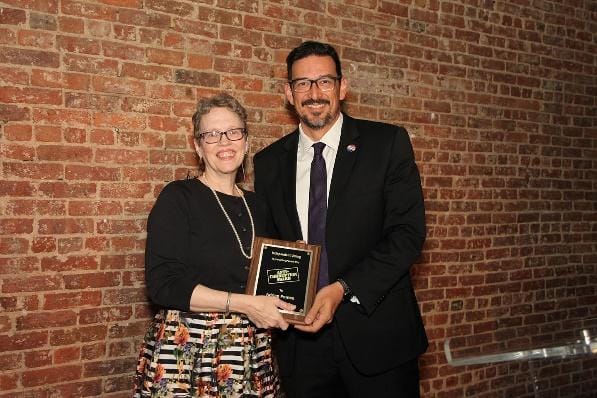 A person in a suit standing in front of a brick buildingDescription automatically generated