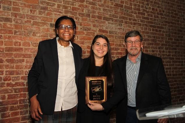 Two people standing in front of a brick wallDescription automatically generated