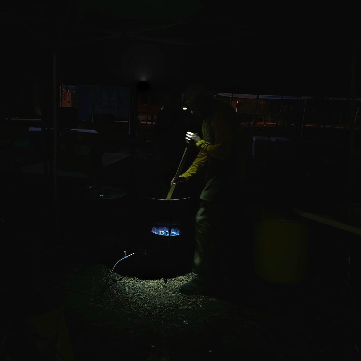 A man wearing a headlamp uses a large oar-like tool to stir a large pot set over a blue flame in an otherwise dark image, taken before dawn.