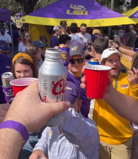 A crowd of people raise their drinks--beer cans and plastic cups--in a toast. In the center of the image, an extremely handsome man who looks like a great writer smiles.