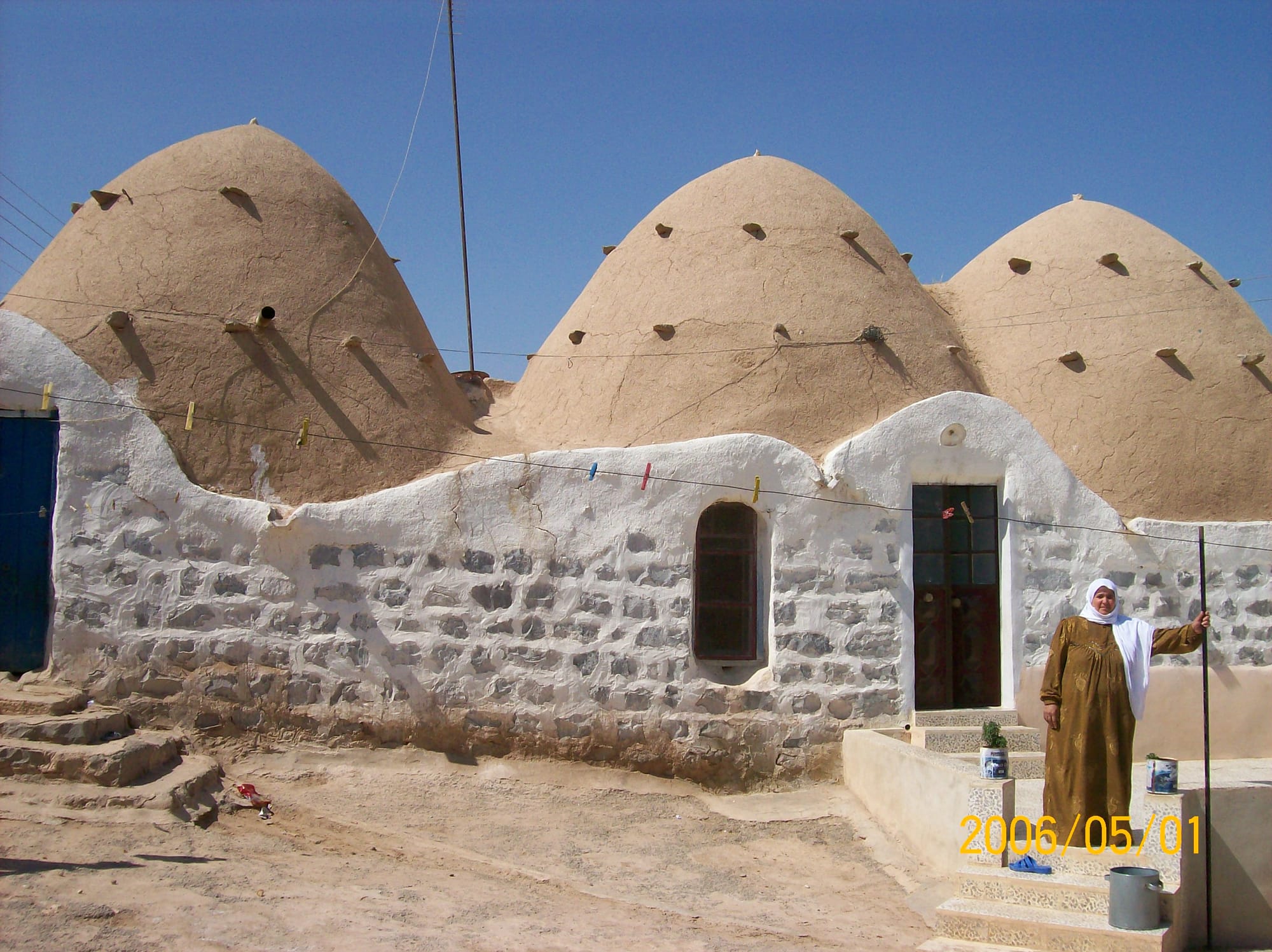A mud and brick three-domed home with a woman on the porch. The woman wears a brown and gold abaya and a white hijab.