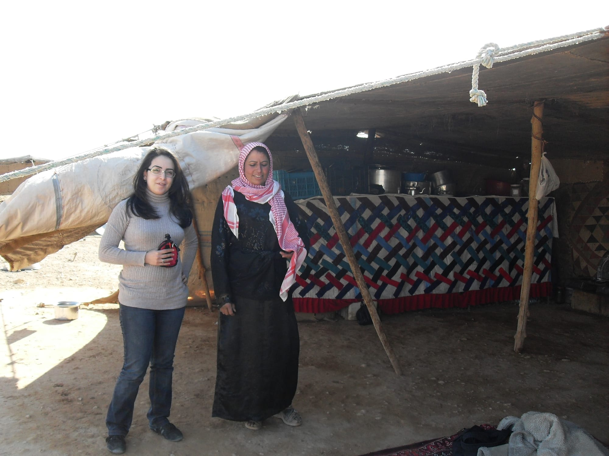 Kindah wearing a sweater and jeans stands next to a Syrian woman wearing a black abaya and red-and-white keffiyah. There are in a large tent. Cooking implements are visible the background.