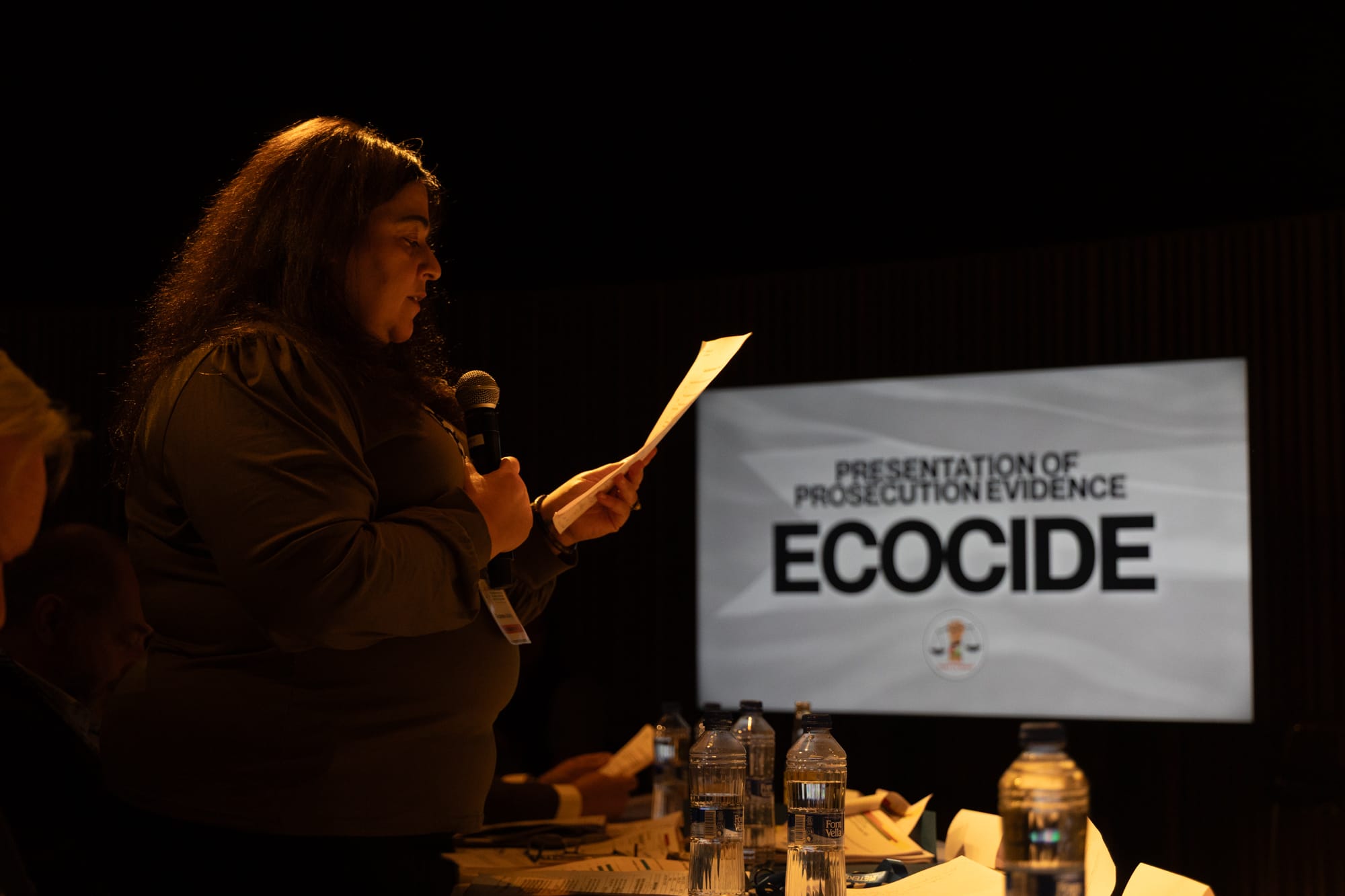 A person stands in a dark room, holding a mic and reaing from a piece of paper. A screen behind them reads "Presentation of prosecution evidence: Ecocide."