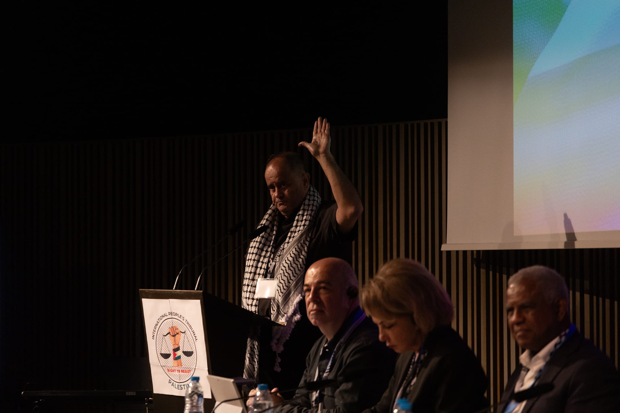 A man wearing a keffiyeh stands behind a podium with his left hand raised. Three jurors sit in the foreground.