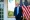 Trump is wearing a dark blue suit with a light blue tie and standing in front of three American flags and a large gold-rimmed mirror.