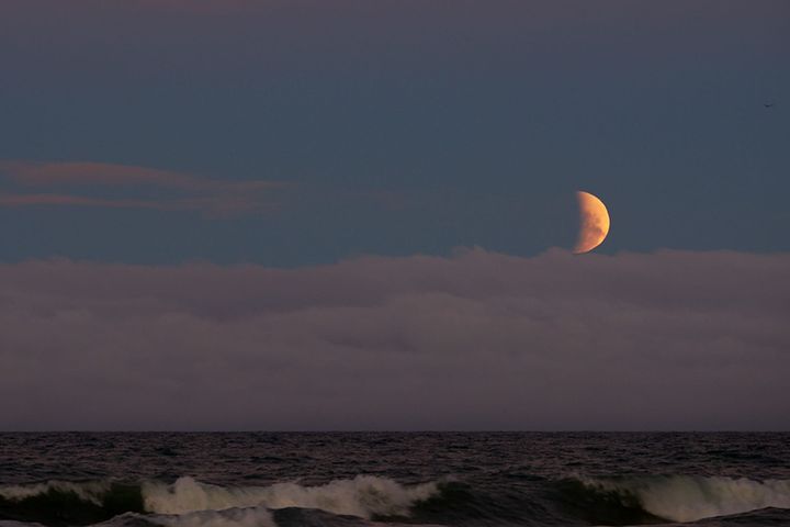 A half moon over clouds on the horizon and the ocean with waves crashing on Waimairi Beach, New Zealand in the foreground. | Photo © 2023 Gary Easterbrook All rights reserved.