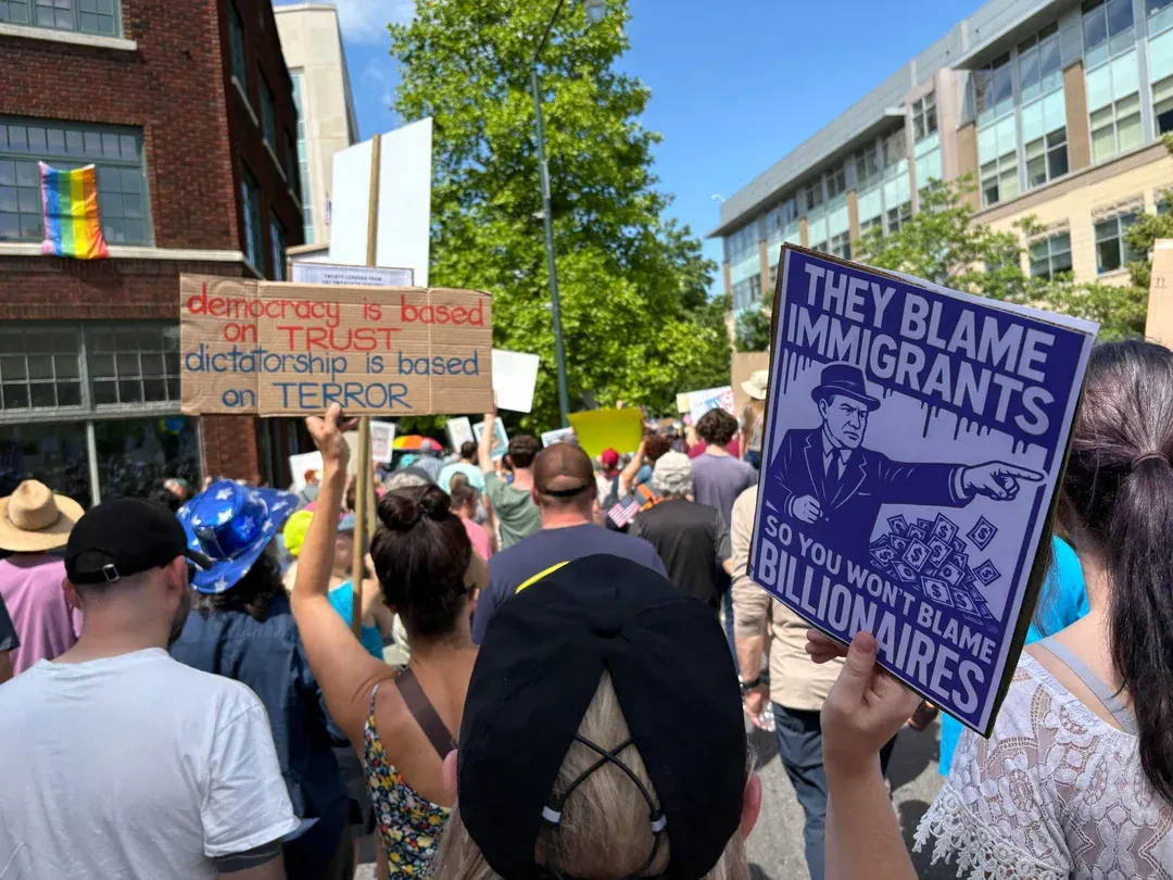 Image from the 'No Kings' rally in Asheville, NC. A crowd of men and women standing in the street holding signs aloft. One reads, "Democracy is based on trust; dictatorship is based on terror.' Another reads, "They blame immigrants so you won't blame billionaires."