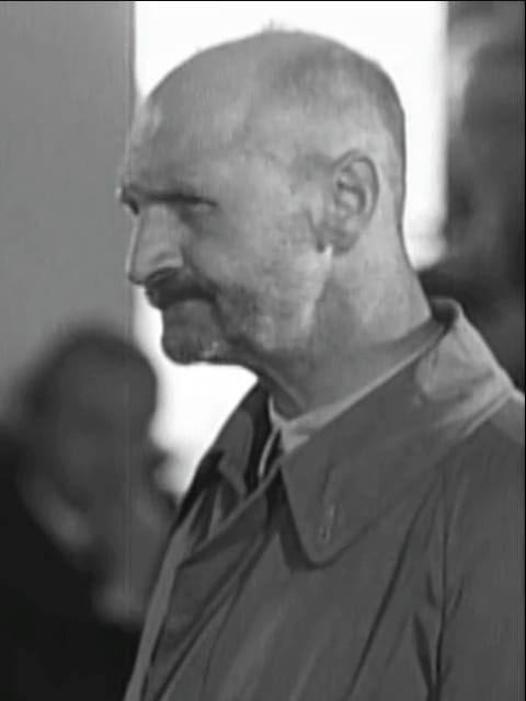 A black-and-white photo of a white man with a shaved head and small moustache staring grimly ahead.