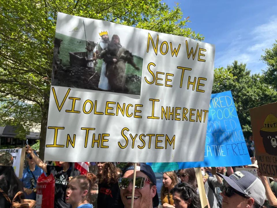 A man at a public demonstration holds a sign. The text reads, "Now We See the Violence Inherent in the System."