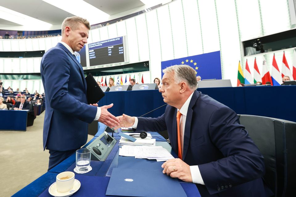 Two men dressed in blue suits shake hands across a table draped with blue cloth. The man on the left is standing in front of the table and he shakes hands with the other man, seated behind it