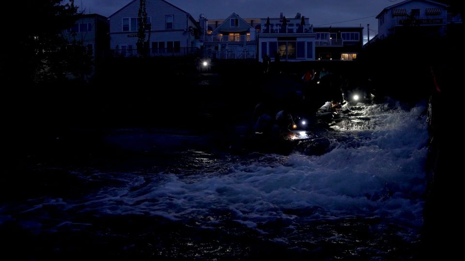 dusk by dam outlet at river, wide image showing buildings in background and people on rocks by spillway with flashlights looking at water.