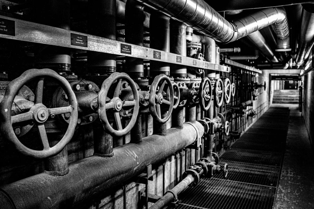 Monochrome photograph of large industrial valves and pipework in a dark corridor, all charcoal metal and low light.