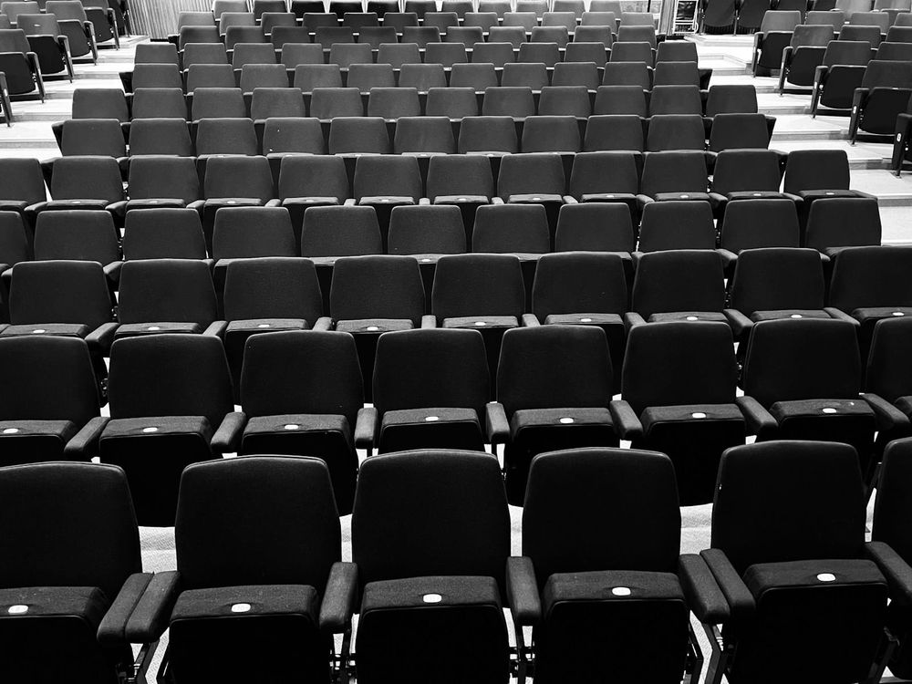 Rows of empty black chairs stretch in a symmetrical grid across a large neutral-toned assembly hall with no people present.