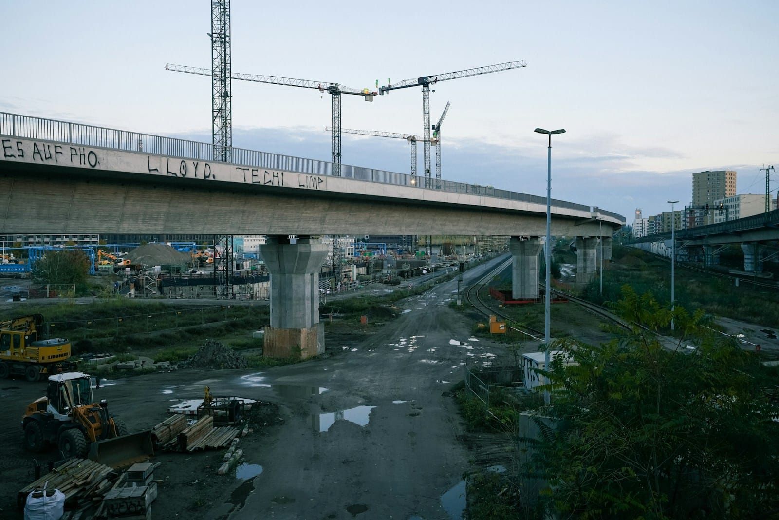 An active construction site with several tall cranes working beneath an overhead bridge span.