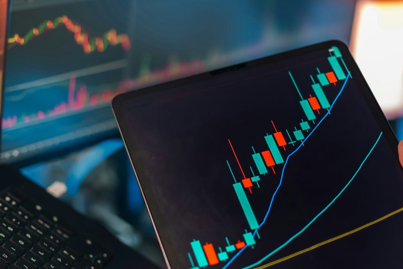 A trading desk with candlestick charts and market data running across multiple screens and a tablet.