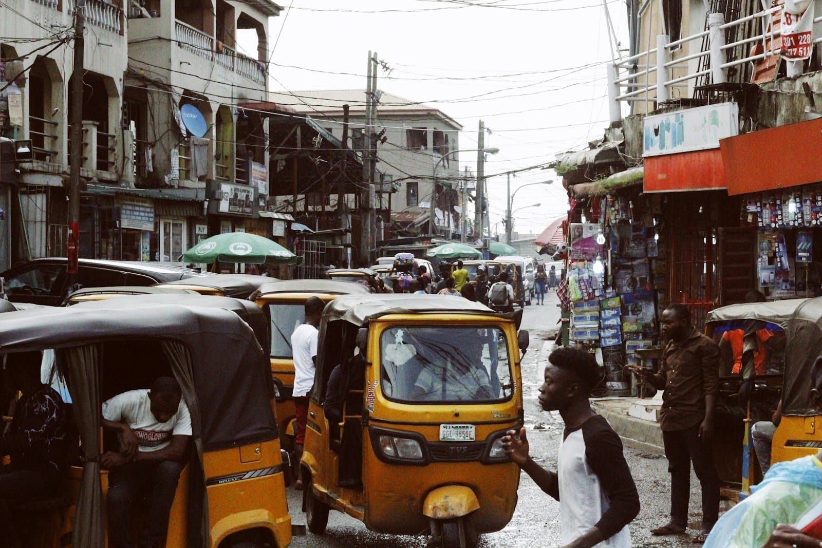 A dense, busy street in Lagos crowded with pedestrians and vehicles between buildings in daylight.