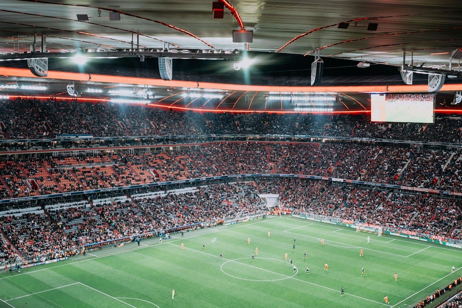 A packed football stadium at night, floodlit pitch and full stands under the dark evening sky.
