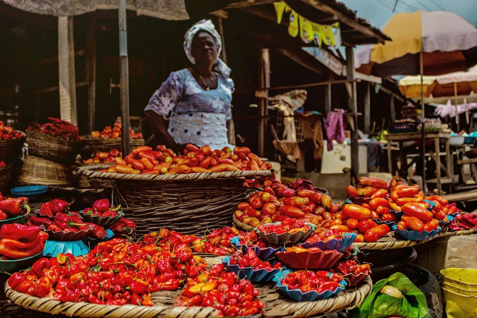 A vendor in white attire standing at his produce stand selling peppers in a market in Abeokuta, Nigeria.