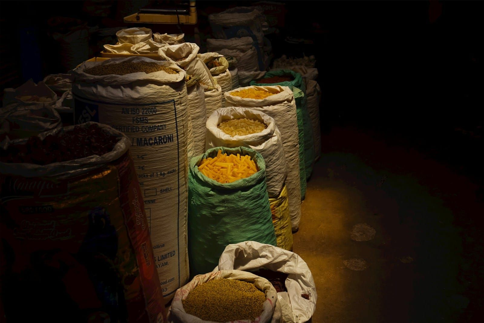 Burlap sacks of grain stacked in a dimly lit warehouse storage area in warm, moody light.
