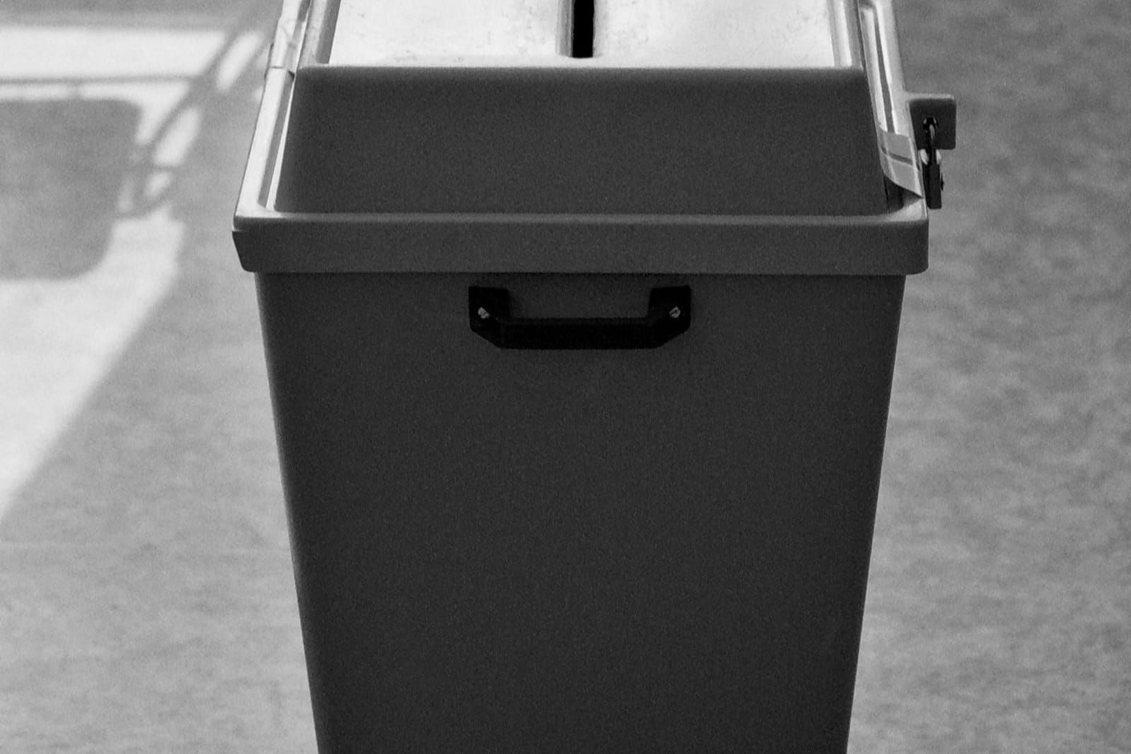 Black-and-white photograph of a plain metal box with a narrow slot, shot straight on with soft shadow and no people.