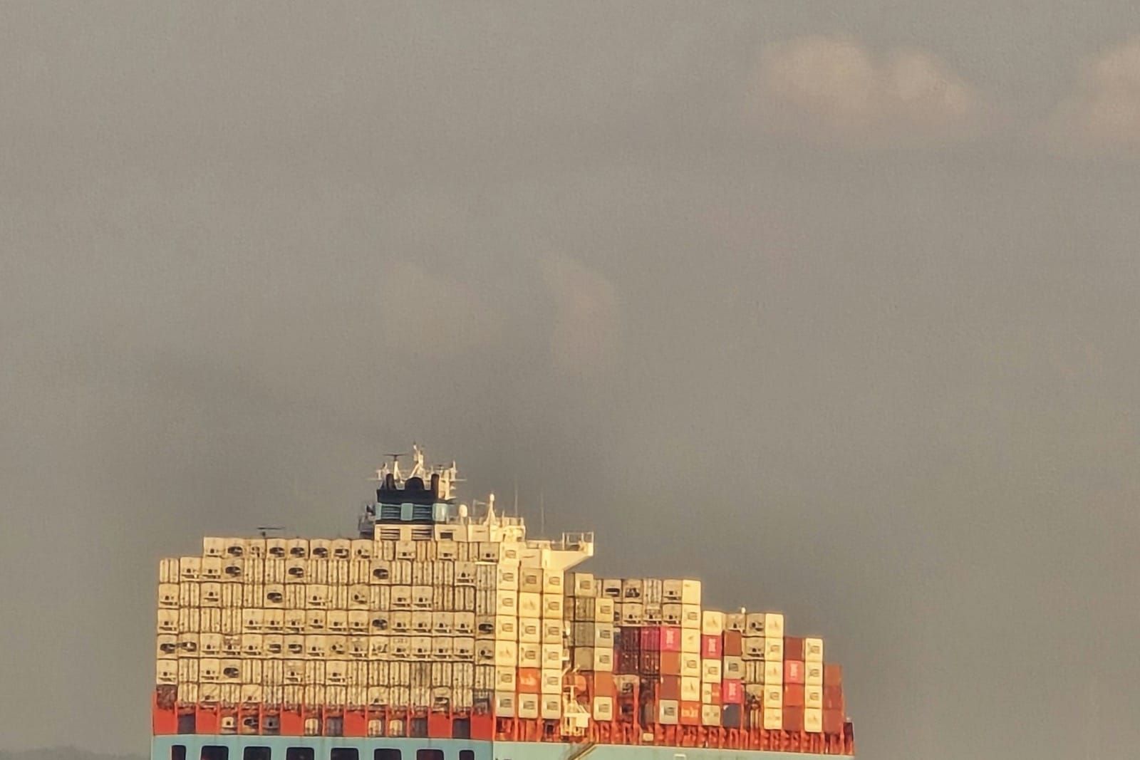 A large loaded container ship sailing across open ocean under a dusk sky.