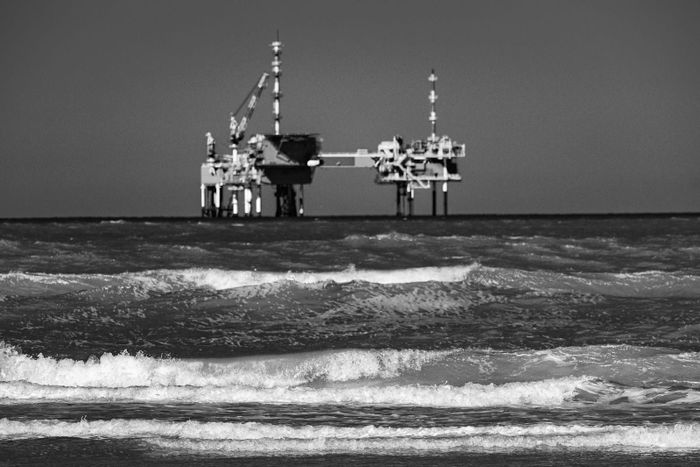 An offshore oil rig platform stands in rough open sea, rendered in desaturated black-and-white tones with the structure dark against a pale sky.