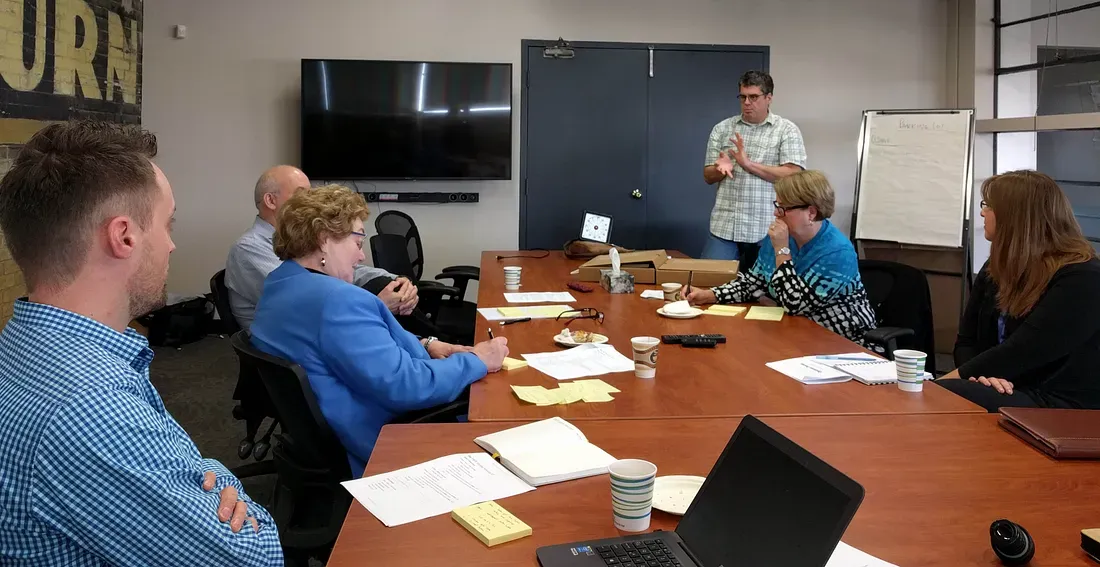 A mixed group of people sitting around a board room table cluttered with papers, pens, and laptops. A man stands at the front of the room, talking.