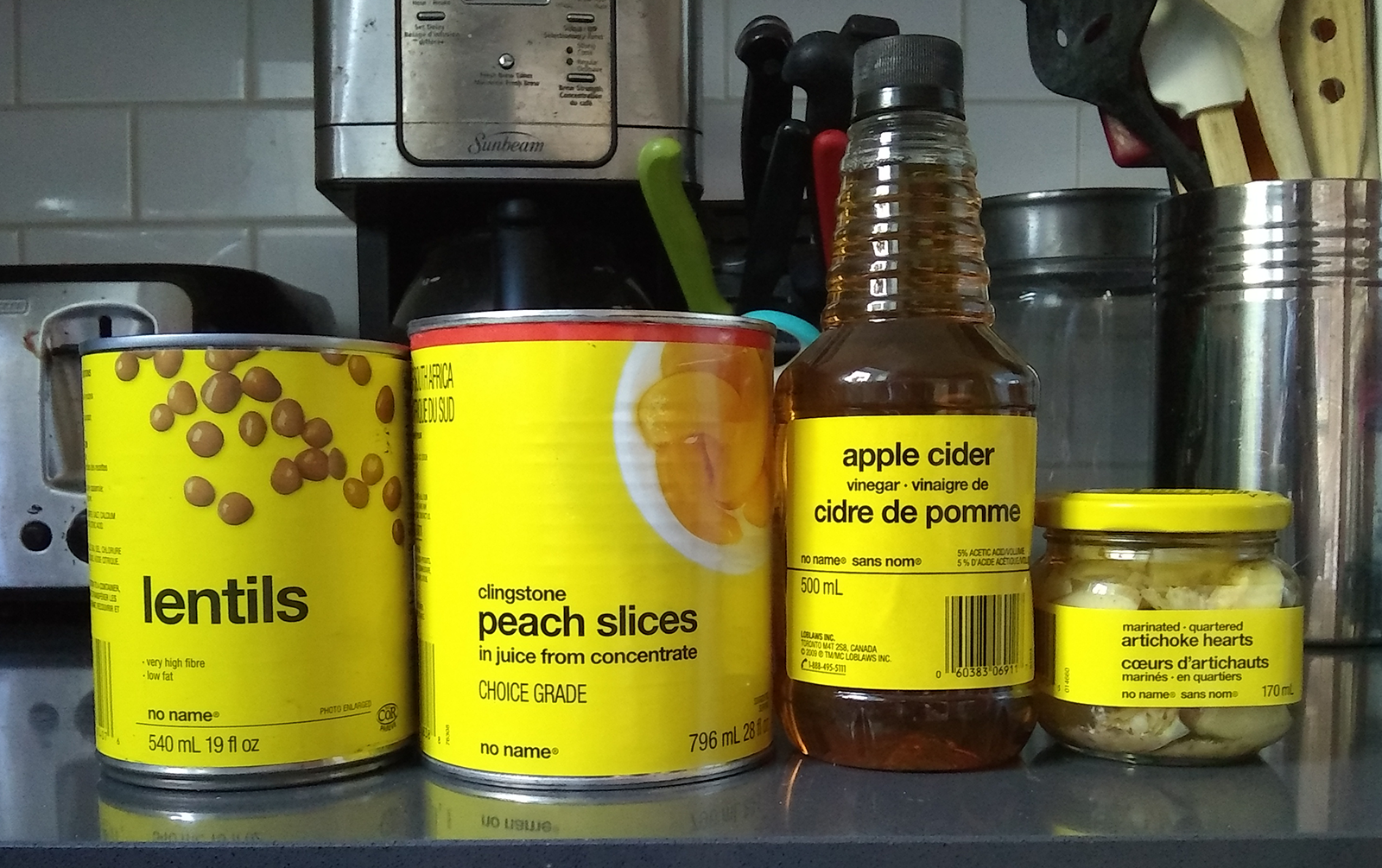No Name brand containers of lentils and peach slices, a bottle of apple cider and a jar of marinated artichoke hearts are lined up on a kitchen counter.