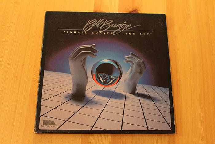 A square video game package on a table. The package has a photo of mannequin hands reaching for a chrome ball.