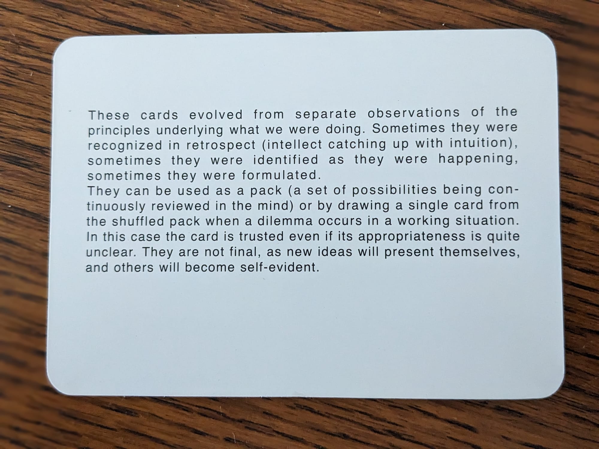 White instruction card with black text sitting on a wooden table. The card describes how to use the Oblique Strategies deck.