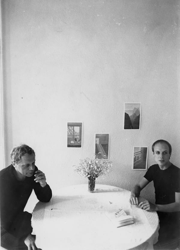 Photo of two men sitting at a table with a vase of flowers and a notebook between them. One is smoking. There are photos on the wall.