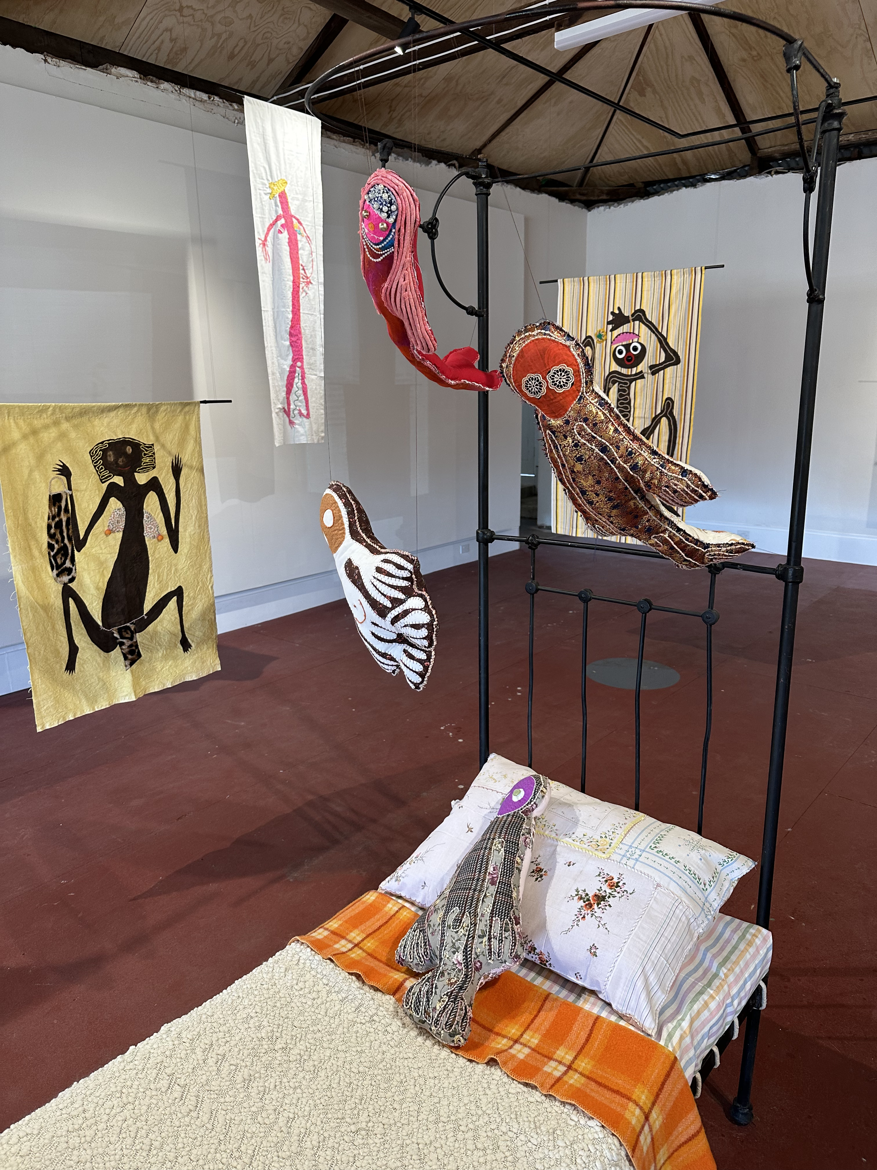 Suspended soft scultpures above a wrought iron bed with previously mentioned artworks in the background