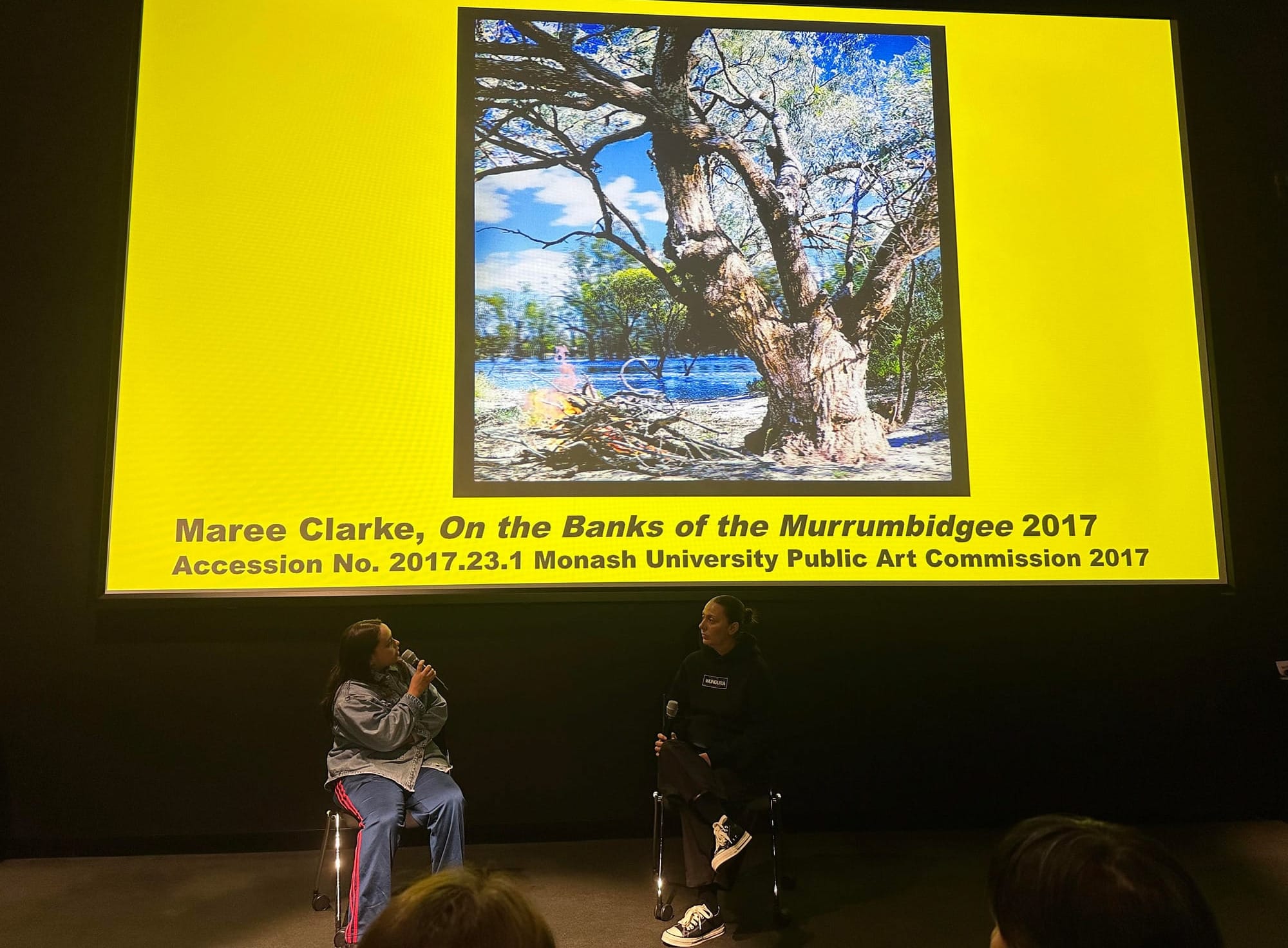 Two women in casual attire sit beneath a photograph of a large and ancient tree on a river bank, they're in deep conversation.