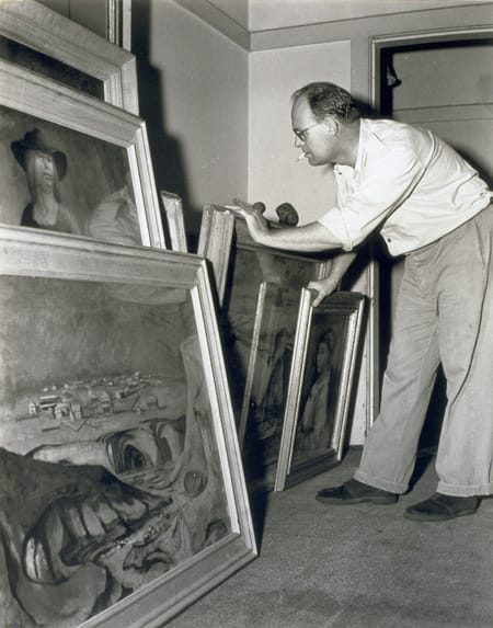 A black and white photograph of a man looking through a stack of framed artworks, cigarette in his mouth.