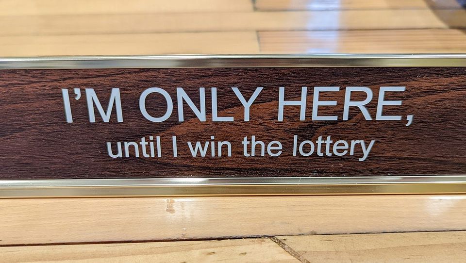 Desk placard reading "I'M ONLY HERE, until I win the lottery". 