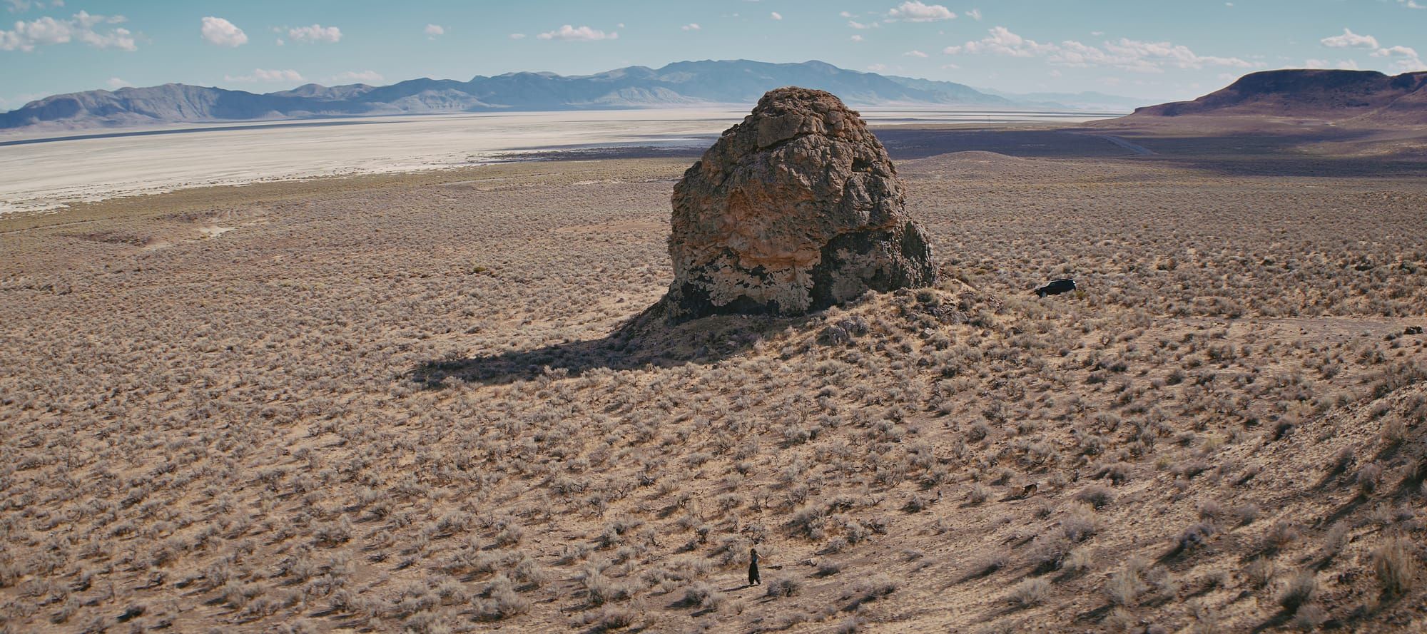 A lone boulder stands on a sagebrush slope above a white playa, with a small figure walking below it.