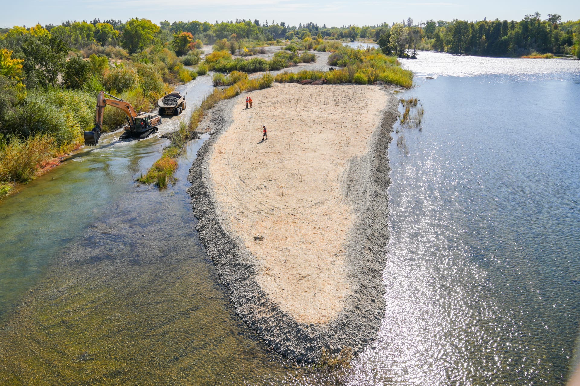 A sandbar on the edge of a river is being restored with heavy machinery visible on the left side of the photo. Trees and shrubs line the river.
