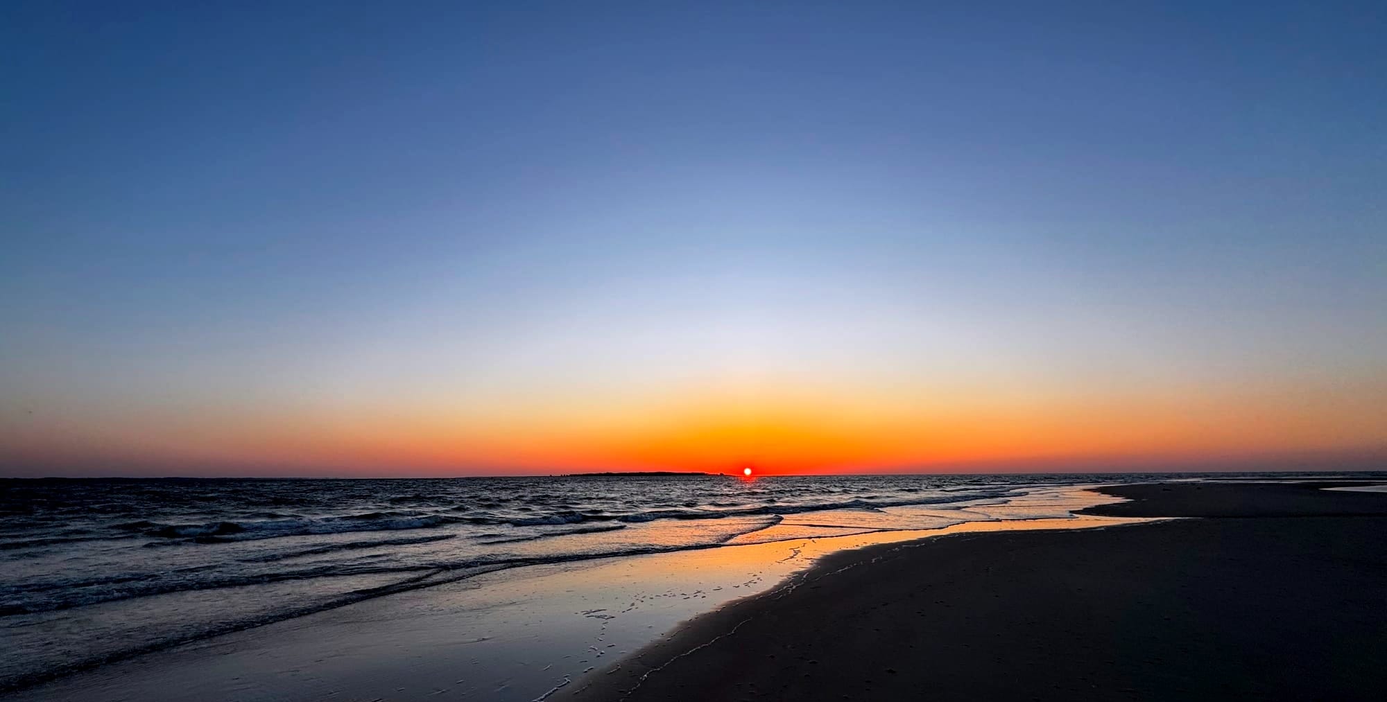 Sun rising over a large body of water with a few small waves, a sandy beach in the foreground, and clear skies colored from red to deep blue overhead.